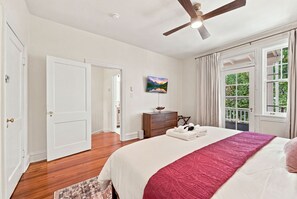 A bedroom with a bed, a ceiling fan, a painting, a window with curtains, and a wooden dresser.