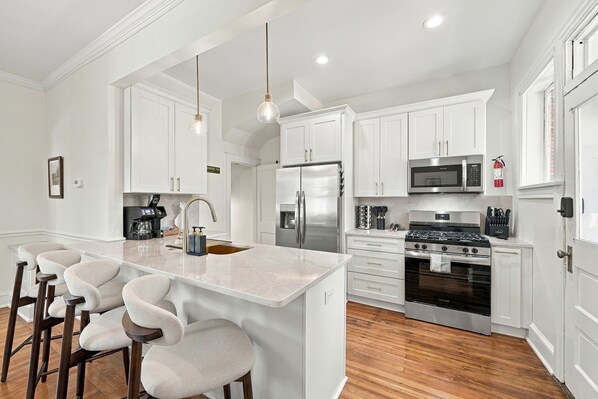 A modern kitchen with white cabinetry, a stainless steel refrigerator, a built-in oven, and a bar area with stools.