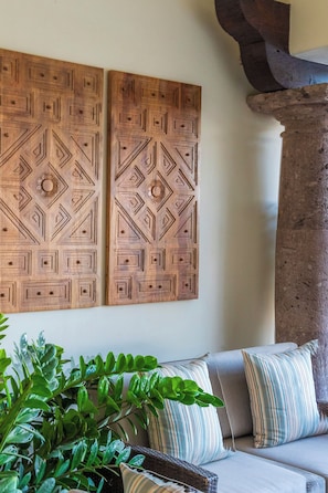 Ein Zimmer mit einer Couch, zwei an der Wand befestigten Holzpaneelen mit geometrischen Mustern, einer großen Topfpflanze und einer Steinsäule.