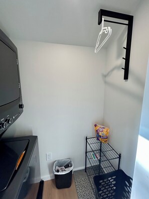 A compact room with a black appliance, a metal rack with a bag of cleaning supplies, a black trash bin, and a patterned rug. - Cozy 2-bedroom cottage between Wildwood & Cape May NJ (Rio Grande)