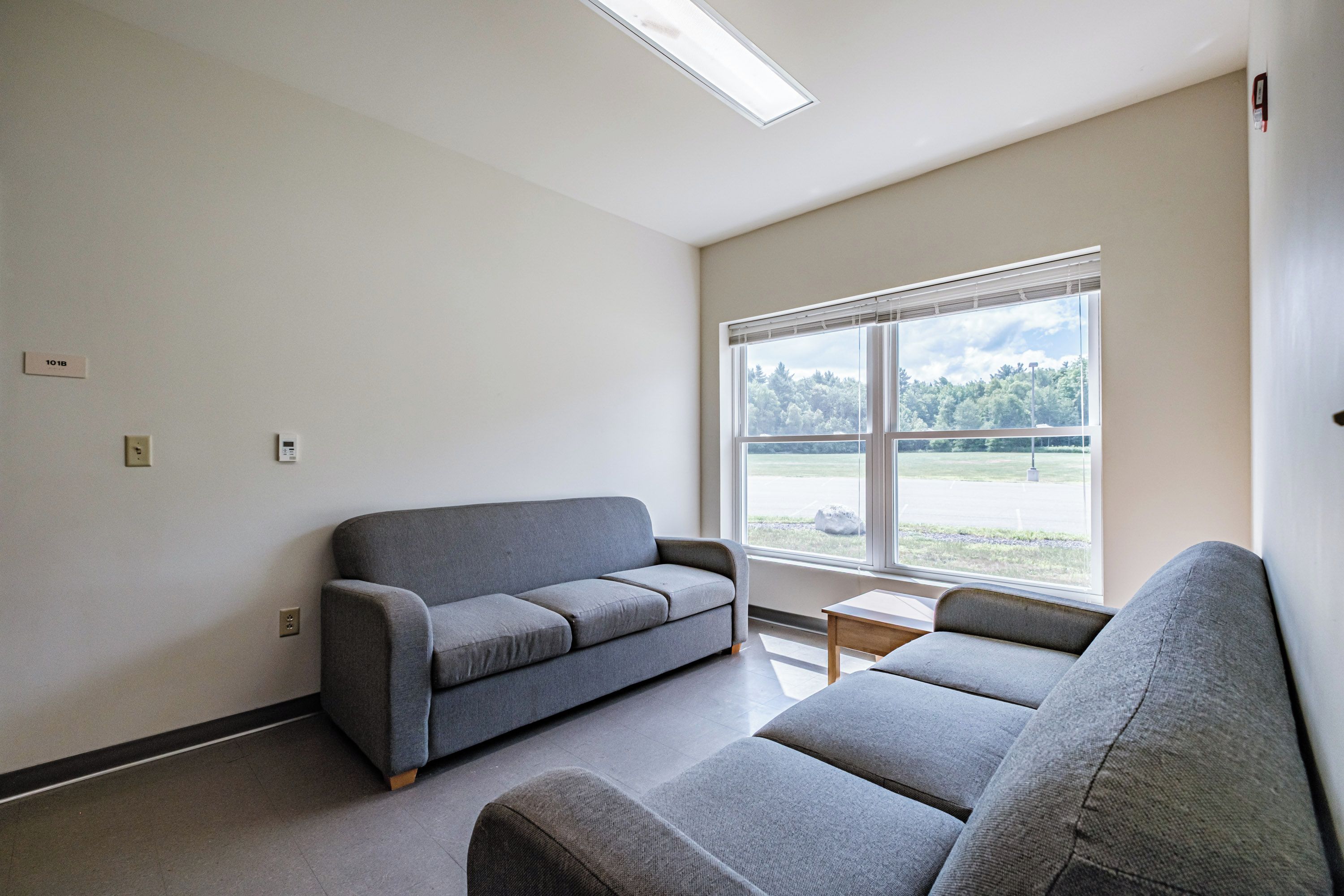 Una habitación con un sofá gris, una mesita y un ventanal con vistas a árboles y un campo.