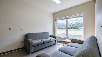 Una habitación con un sofá gris, una mesita y un ventanal con vistas a árboles y un campo.