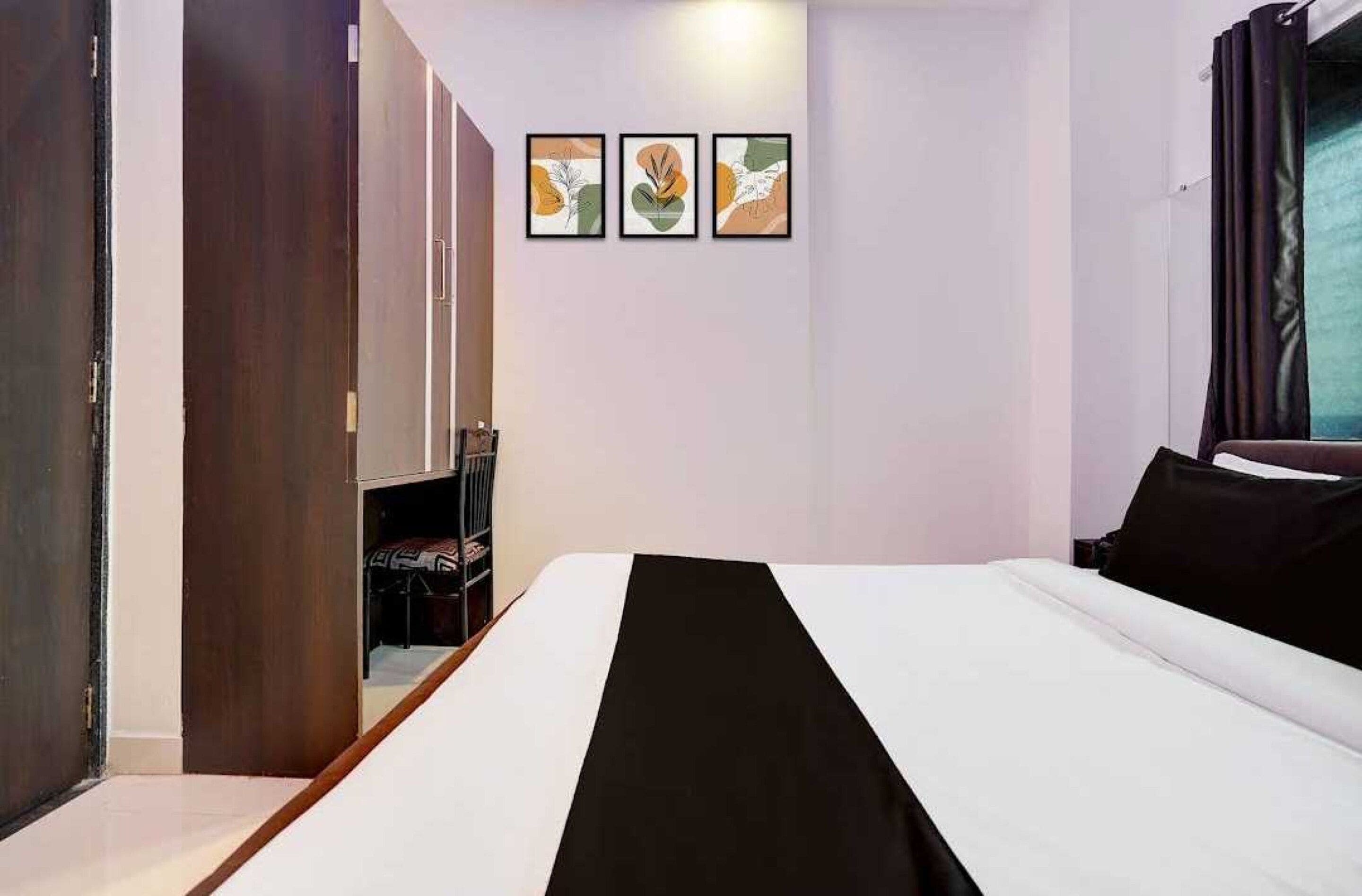 A hotel room with a bed, a wardrobe, and a shelf with books. There are three framed artworks on the wall.