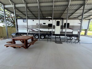 A covered outdoor area with a picnic table and chairs, a grill, and a large RV parked under a metal roof.