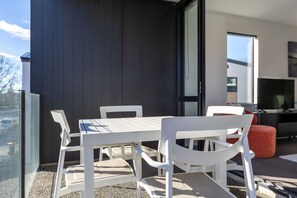 An outdoor balcony with a white table and chairs, a television, and a modern building in the background. - Heart of CHCH - Modern Apt Near Stadium + Parking (Christchurch)