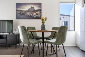 En matplats med ett runt bord, fyra stolar, en vas med blommor, en tavla på väggen, en tv och ett kylskåp.