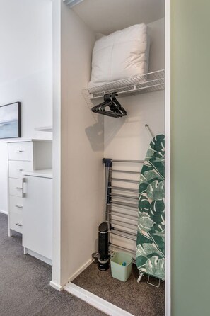 A compact room with a white wardrobe, a drying rack, a heater, and a green patterned item on the floor. - Heart of CHCH - Modern Apt Near Stadium + Parking (Christchurch)