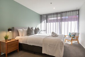 A modern bedroom with a bed, bedside table, lamp, chair, and large window with curtains. - Heart of CHCH - Modern Apt Near Stadium + Parking (Christchurch)