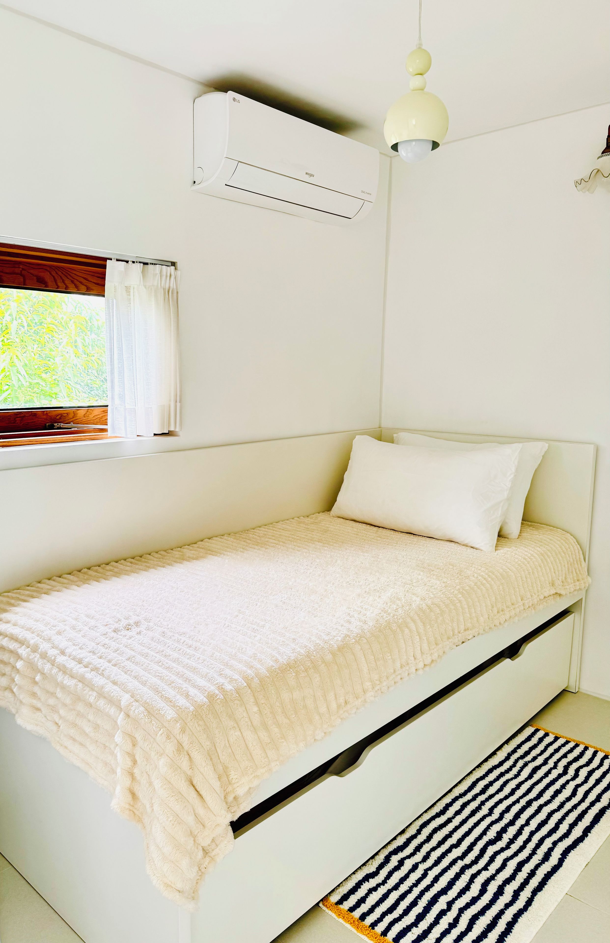 Ein Einzelbett mit weißer Matratze und Kissen, eine weiße Decke, ein Fenster mit Vorhängen, eine Klimaanlage und ein Deckenventilator.