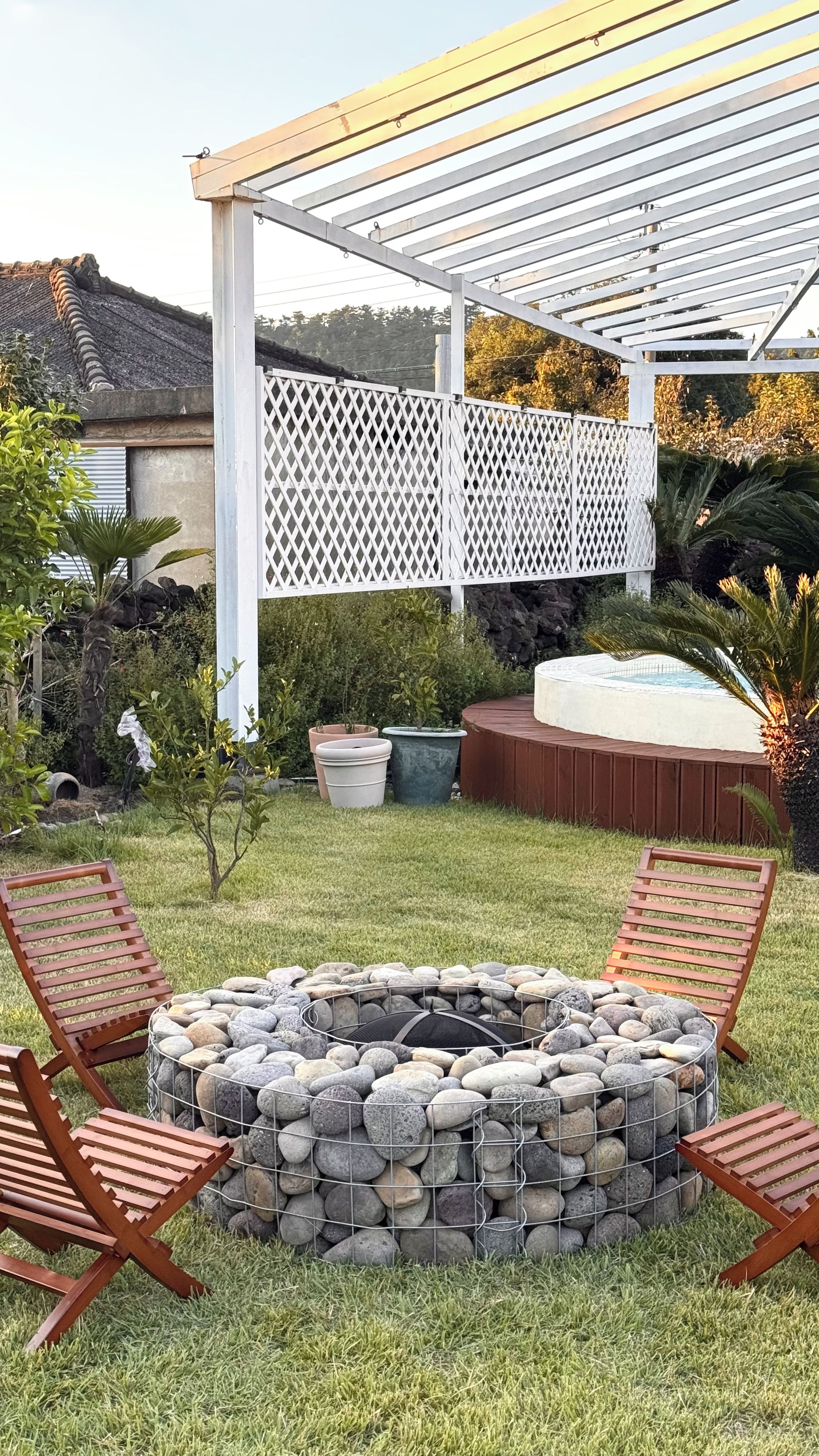 Ein Garten mit einem Steinkamin, zwei Holzliegen, einer weißen Pergola und einem Whirlpool.
