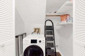 A compact laundry room with a front-loading washing machine, a metal step ladder, and a white shelf with items. - New! Luxury Spacious Home 4 Beds 2 Bath 20 Min DTX (Balch Springs)