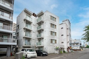 Ein mehrstöckiges Wohngebäude mit Balkonen, ein Parkplatz mit einem weißen und einem schwarzen Auto und einem wolkenlosen Himmel.