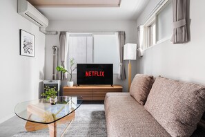 Ein modernes Wohnzimmer mit einem Flachbildfernseher, der 'NETFLIX' anzeigt, einem Glastisch, einer Couch und einem Topf mit Pflanze.