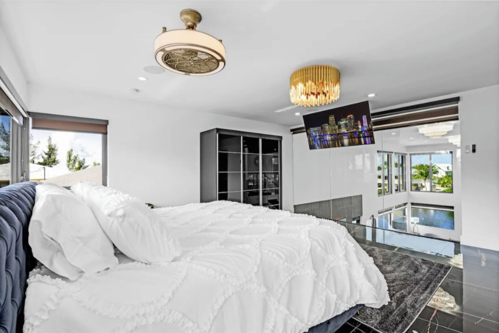 A modern bedroom with a large bed, a ceiling fan, a TV, and a balcony with a pool view.