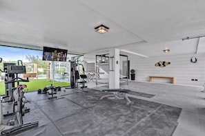 A modern home gym with exercise equipment, a large TV, and a view of a green lawn.