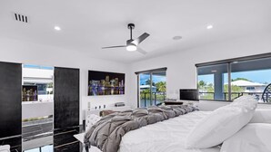 A modern bedroom with a large bed, a ceiling fan, a cityscape view, and a balcony with a view of the city and palm trees. - Heated Pool • Hot Tub • Theater • Waterfront (Key Largo)