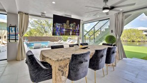 A modern dining area with a large table, comfortable chairs, a flat-screen TV, and a view of a pool and greenery. - Heated Pool • Hot Tub • Theater • Waterfront (Key Largo)