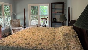 Ein Schlafzimmer mit einem großen Bett, einem Sessel, einem Bücherregal und einem Fenster mit Blick auf Bäume.