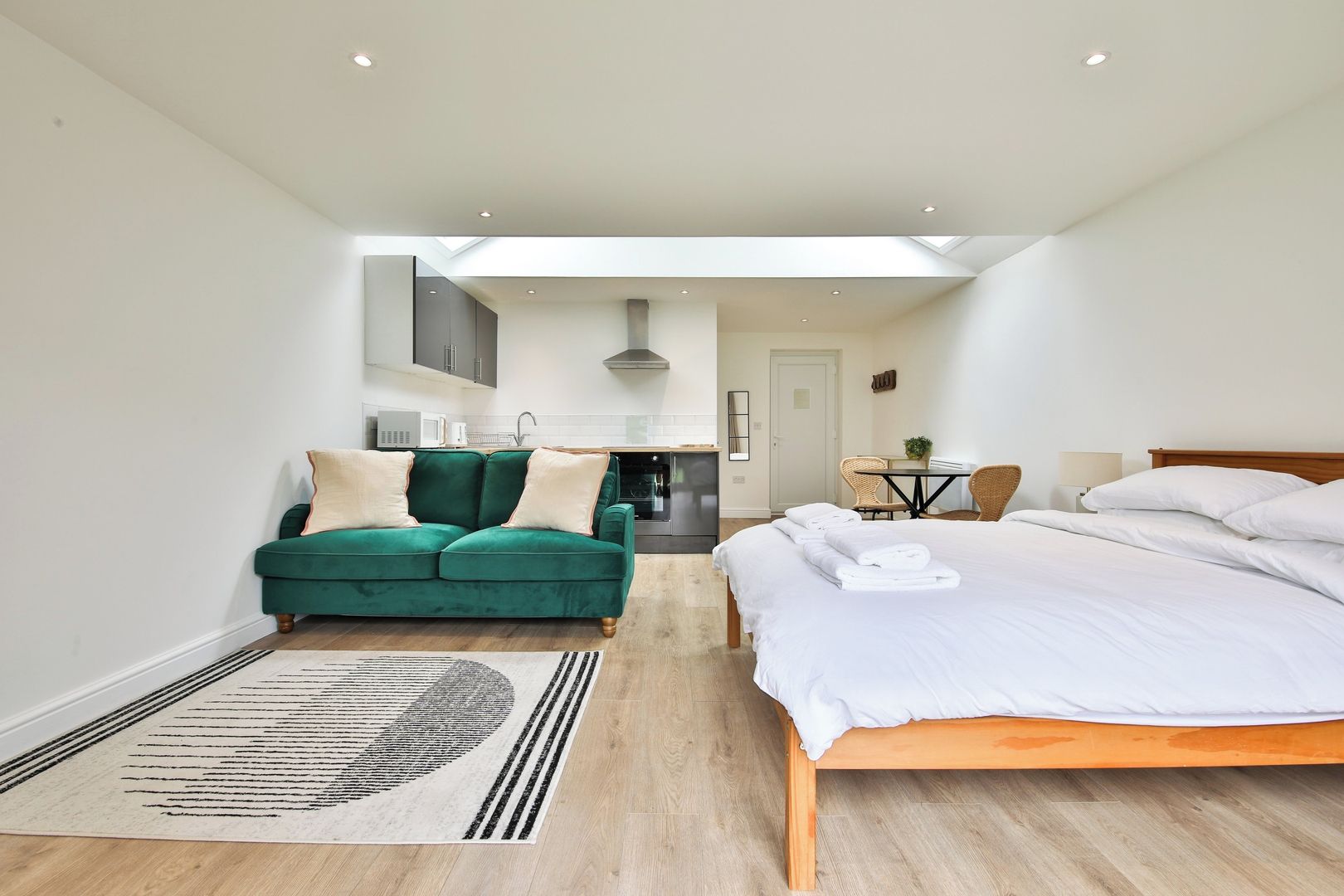 A modern hotel room with a large bed, a green sofa, a dining table, and a kitchen area.