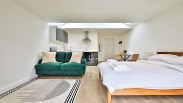 A modern hotel room with a large bed, a green sofa, a dining table, and a kitchen area.