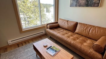 A living room with a brown leather sofa, a wooden coffee table with a magazine and remote, a large window with a view of trees, and a painting on the wall.