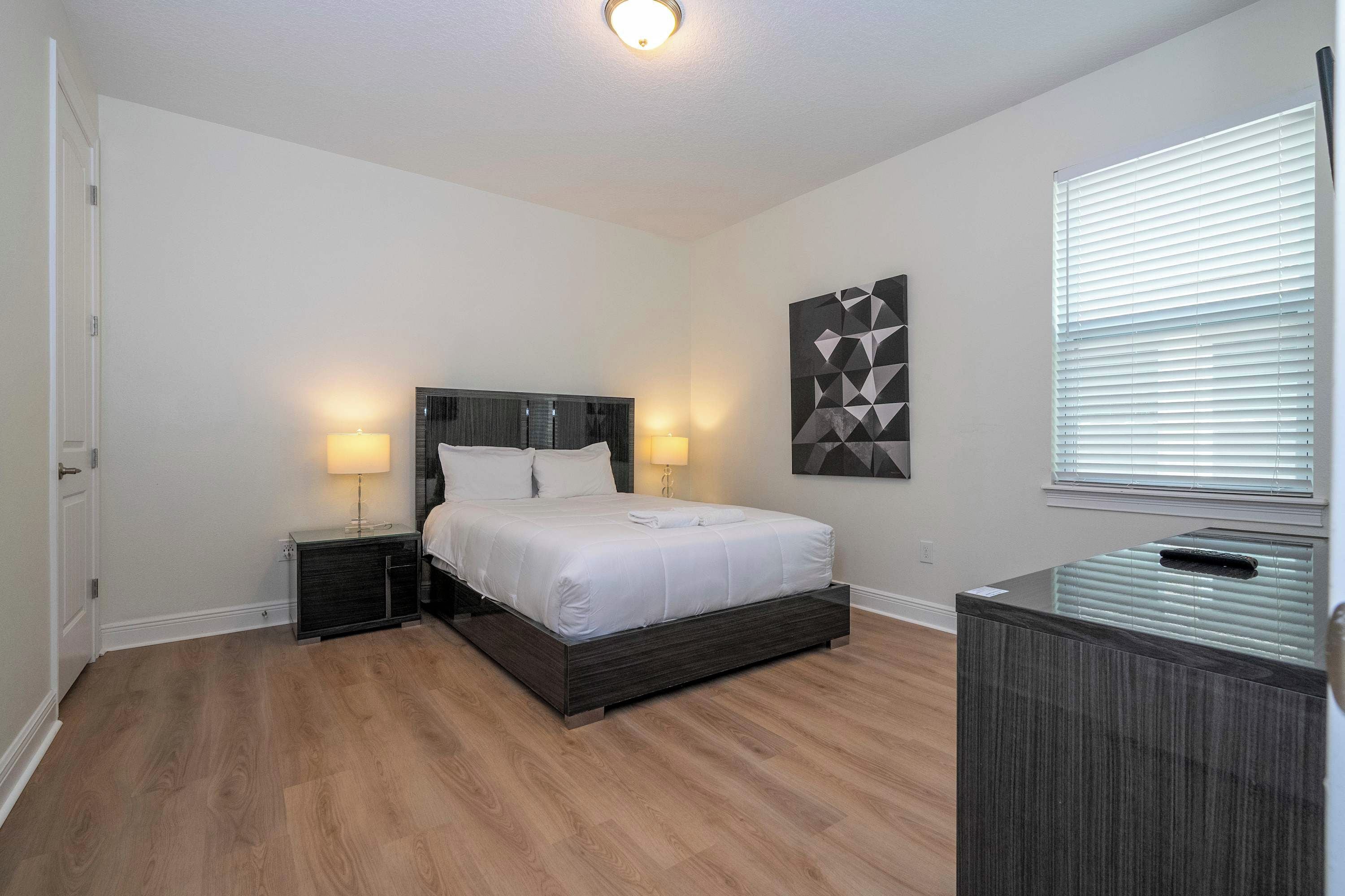 A bedroom with a bed, two bedside tables, a wall-mounted artwork, and a window with blinds.