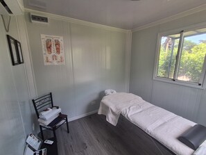 A room with a single bed, a small table with a chair, a poster on the wall, and a window with a view of greenery.