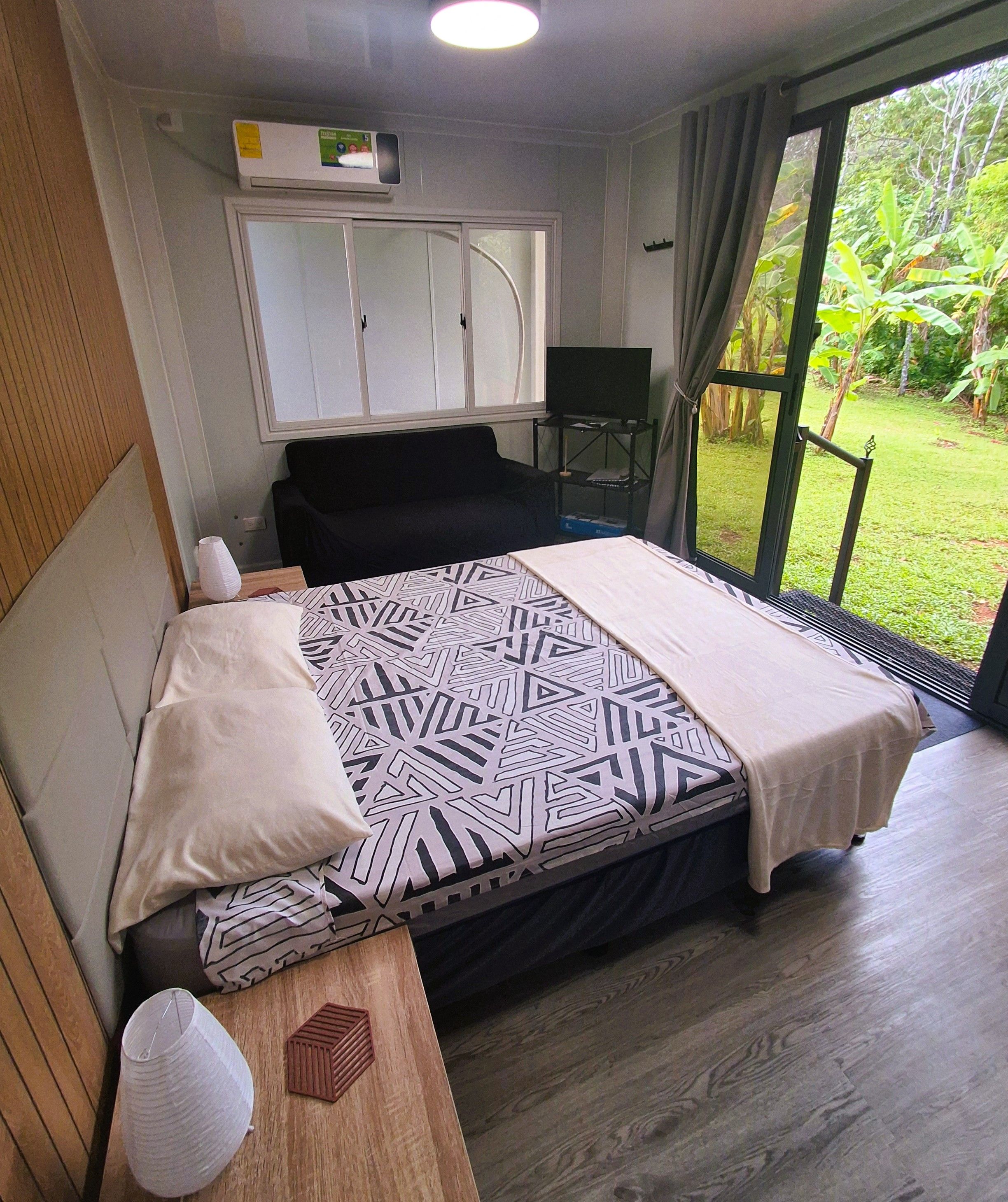 A bedroom with a bed, a desk, a chair, and a view of a garden.