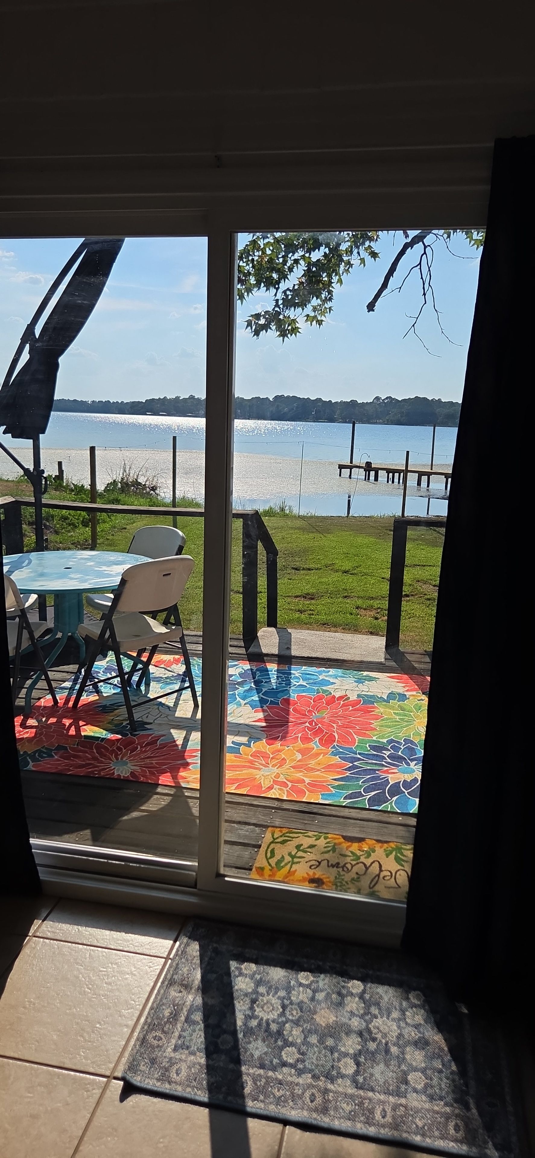 A patio area with a table and chairs, a colorful rug, and a view of a lake through a glass door.