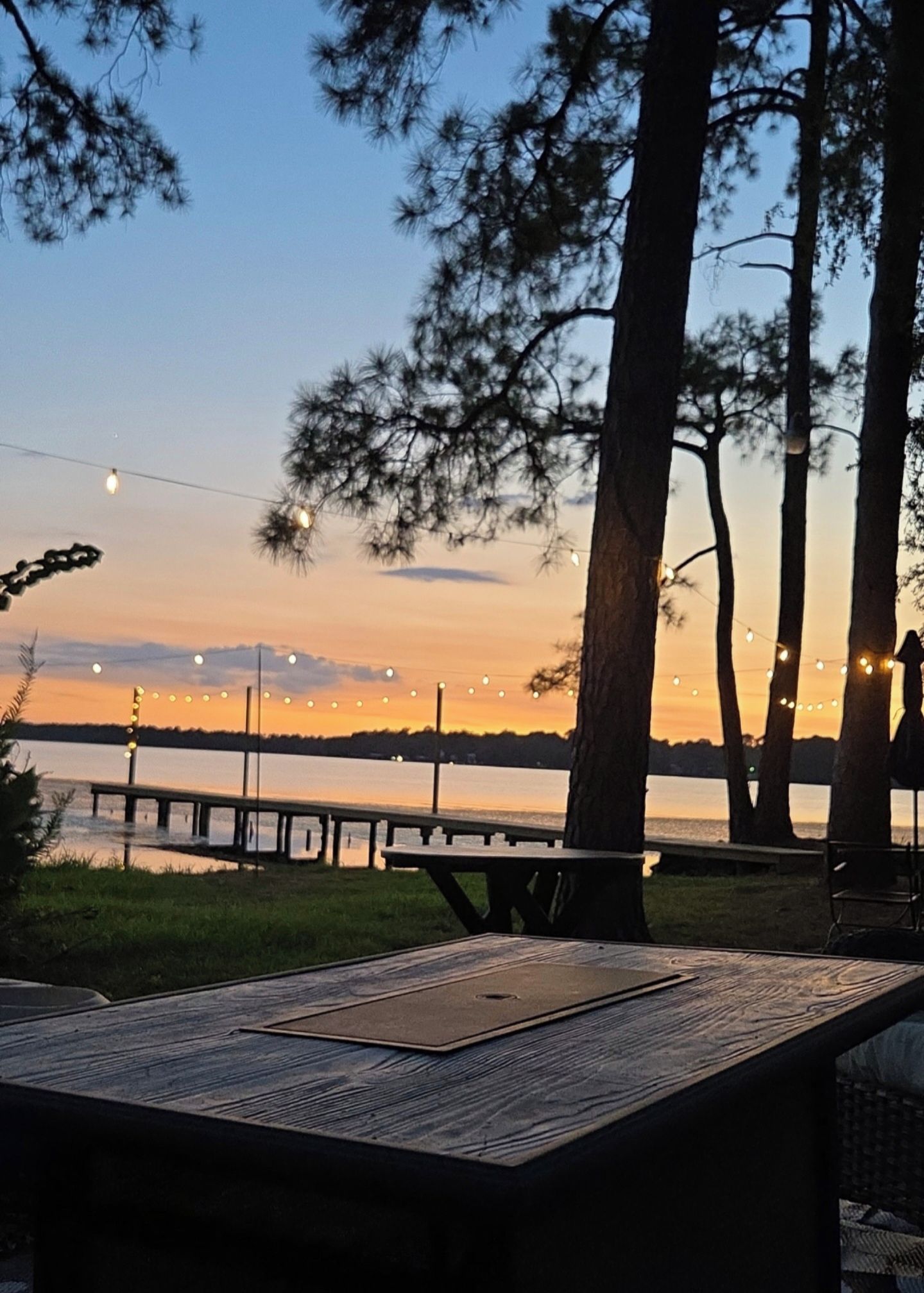 A picnic table with a view of a sunset over a body of water, surrounded by trees and string lights.