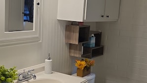 Ein Badezimmer mit weißem Waschbecken, Spiegel und Schrank, einer Toilette und einem kleinen Ablagefach für Toilettenartikel.