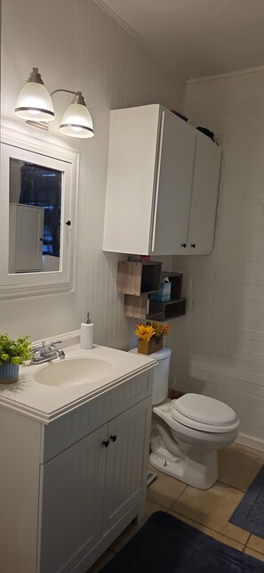 A bathroom with a white sink, mirror, and cabinet, a toilet, and a small shelf with toiletries.