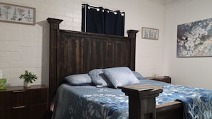 Ein Schlafzimmer mit einem Holz-Kopfteil, einem Bett mit blumigem, blauem Bettbezug, einem Nachttisch mit einer Pflanze und einem Bild an der Wand.