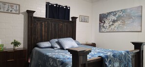 A bedroom with a wooden headboard, a bed with a blue floral bedspread, a nightstand with a plant, and a painting on the wall.