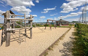 A sandy playground with wooden climbing structures and swings, a covered seating area, and a view of the sea with boats in the background. - Gorgeous home in Juelsminde with WiFi (Juelsminde)
