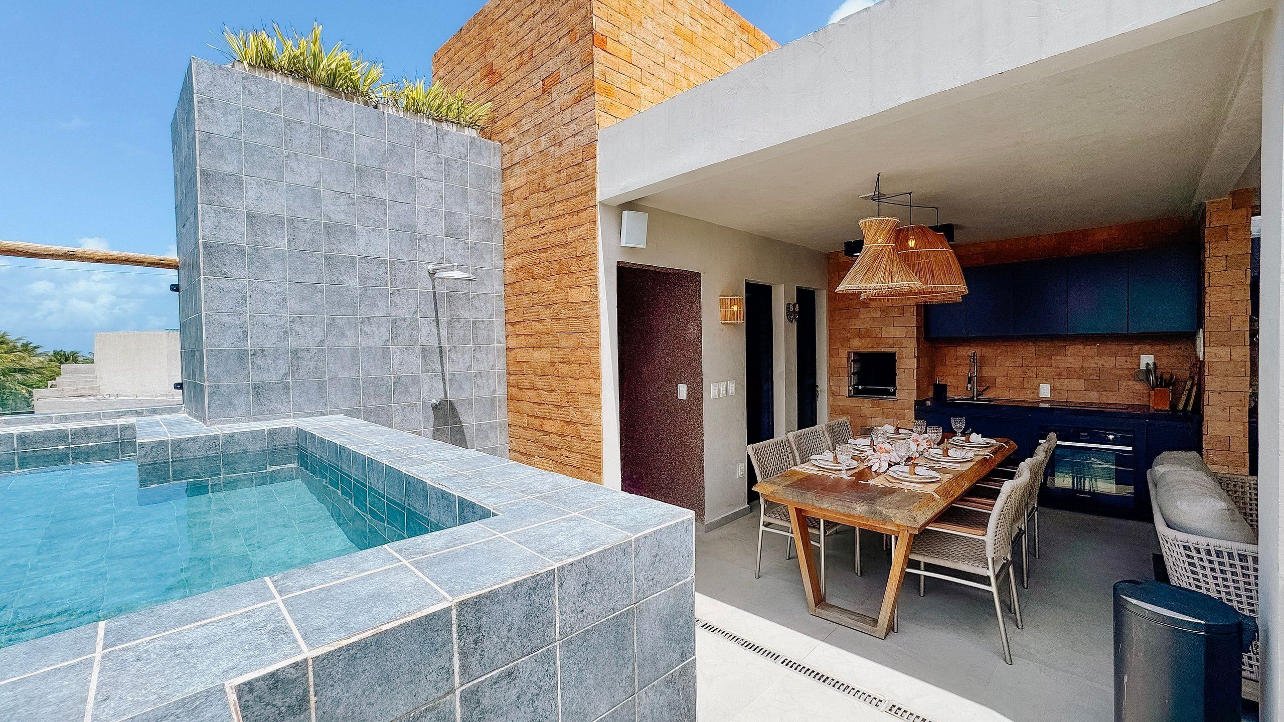 Uma área de piscina moderna ao ar livre, com muro de pedra e espaço de refeições com mesa de madeira e cadeiras de vime.