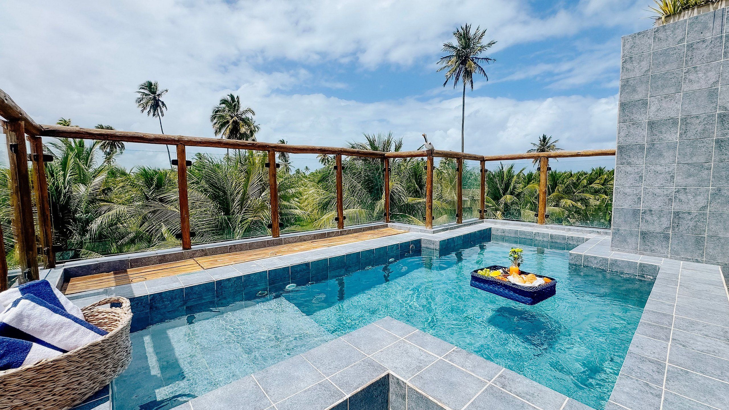 Piscina infinita no terraço com deck de madeira, rodeada por palmeiras e céu claro.