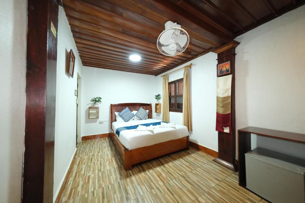 A bedroom with a wooden ceiling, a bed with a white mattress and blue bedding, a bedside table, a wall-mounted fan, and a window with curtains.