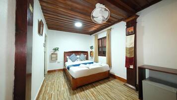 A bedroom with a wooden ceiling, a bed with a white mattress and blue bedding, a bedside table, a wall-mounted fan, and a window with curtains.