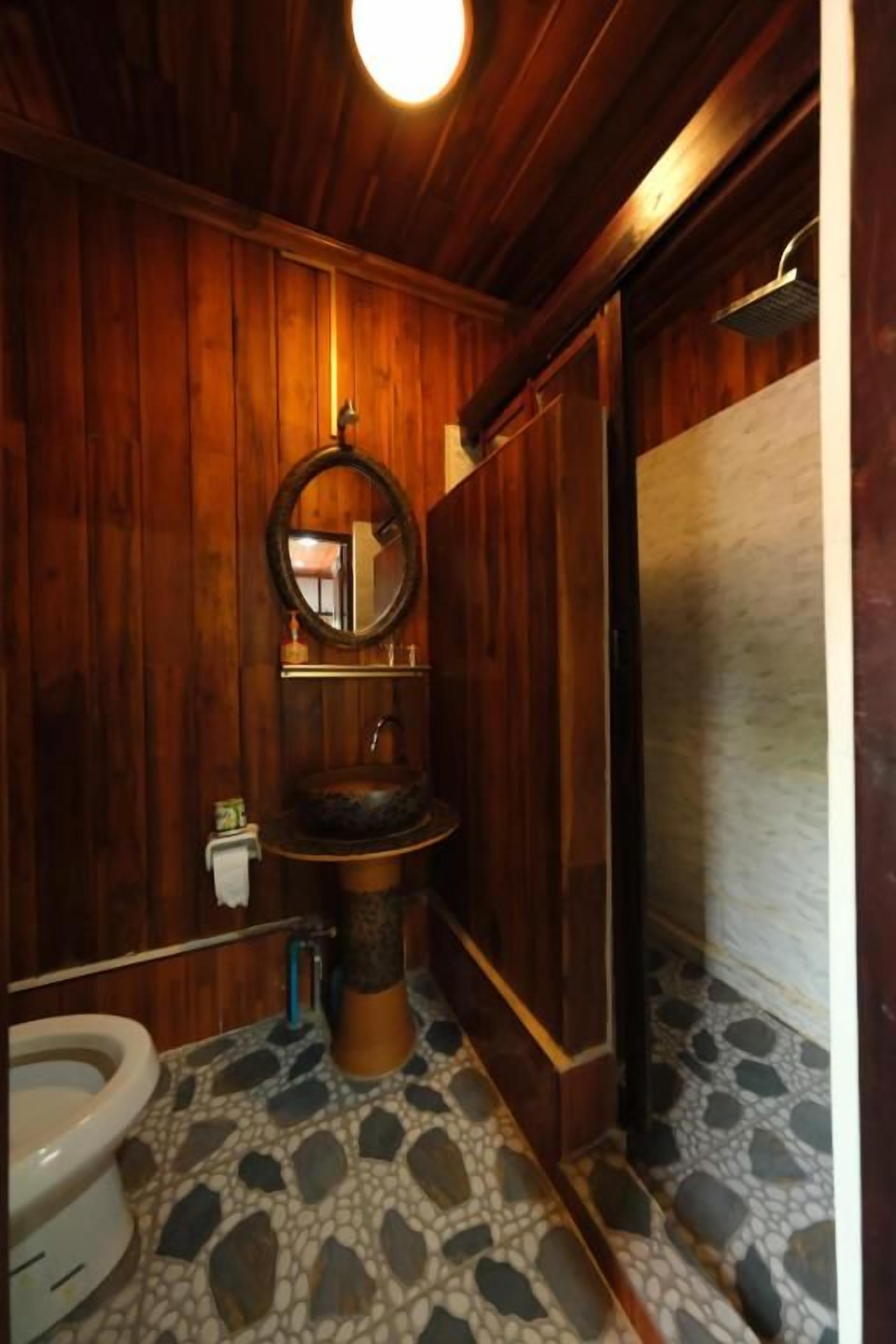 Un baño con paneles de madera, un espejo redondo, un lavamanos sobre pedestal y un inodoro.
