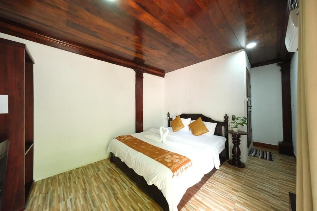A hotel room with a wooden ceiling, a bed with a white comforter and brown pillows, a small bedside table with a plant, and a wooden floor.