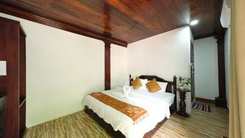 A hotel room with a wooden ceiling, a bed with a white comforter and brown pillows, a small bedside table with a plant, and a wooden floor.