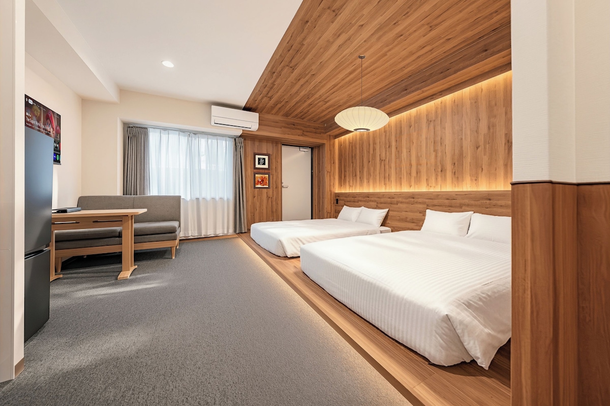 A hotel room with a wooden ceiling, two beds, a desk with a chair, and a large window with curtains.