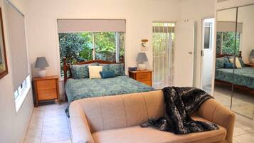 A bedroom with a bed, a sofa, a nightstand, a lamp, and a window with a view of greenery.