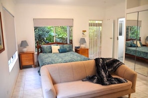 A bedroom with a bed, a sofa, a nightstand, a lamp, and a window with a view of greenery.
