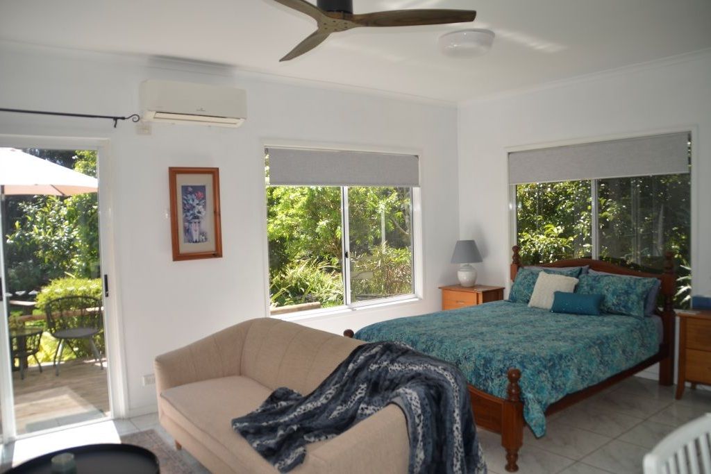 A bedroom with a bed, a sofa, a ceiling fan, and a window with a view of greenery.