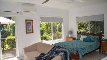 A bedroom with a bed, a sofa, a ceiling fan, and a window with a view of greenery.