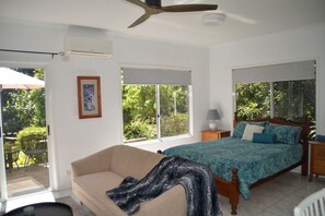 A bedroom with a bed, a sofa, a ceiling fan, and a window with a view of greenery.