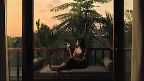 A person sitting on a balcony with a glass of wine, surrounded by lush greenery and a sunset view. - Villa Omah Bumbak (Badung)