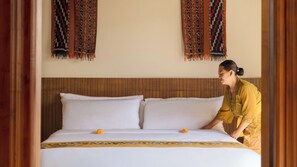 A bed with white linens and a decorative border, a wooden headboard, and a person in traditional attire adjusting the bed. - Villa Omah Bumbak (Badung)